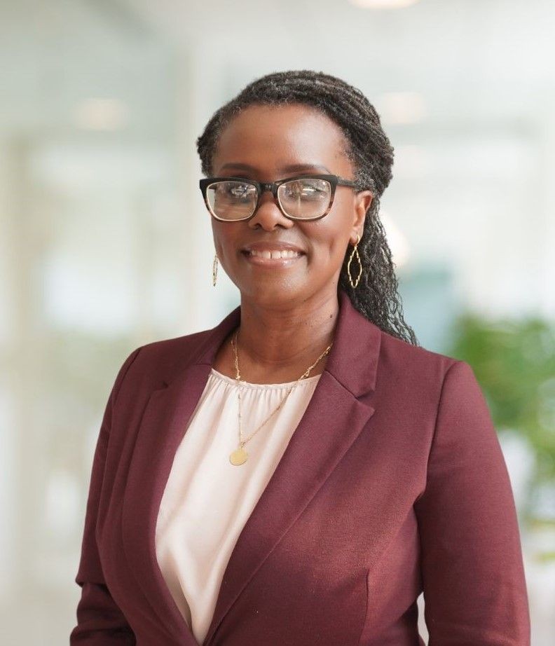 Smiling person in a maroon blazer and glasses standing in a bright room.
